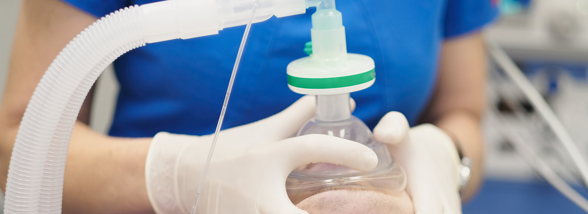 A person in a white lab coat is handling a clear plastic tube connected to a medical device with a green cap, set against a blue background.